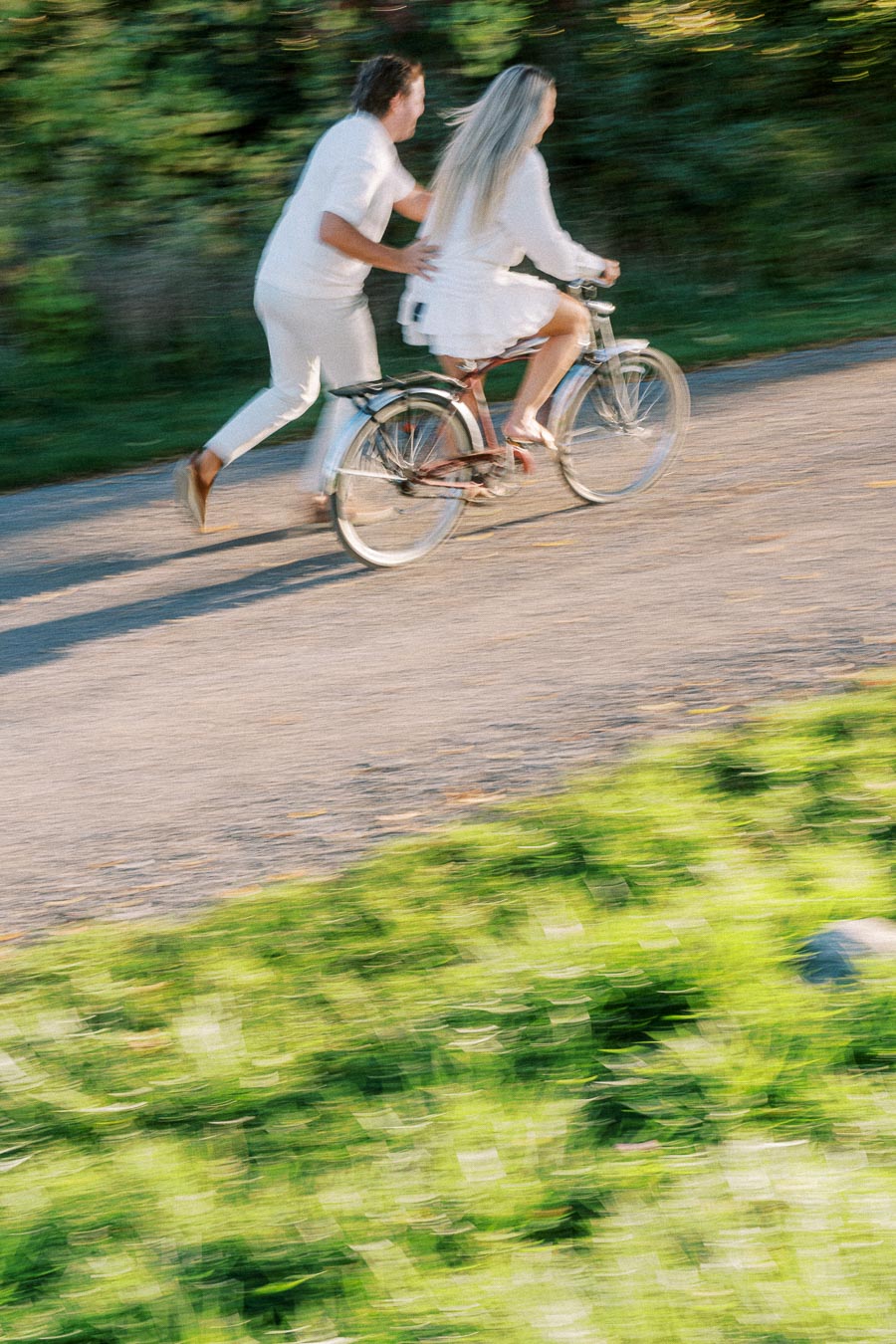 A couple in white outfits biking together on a sunlit path surrounded by greenery, capturing a playful and joyful moment in motion.