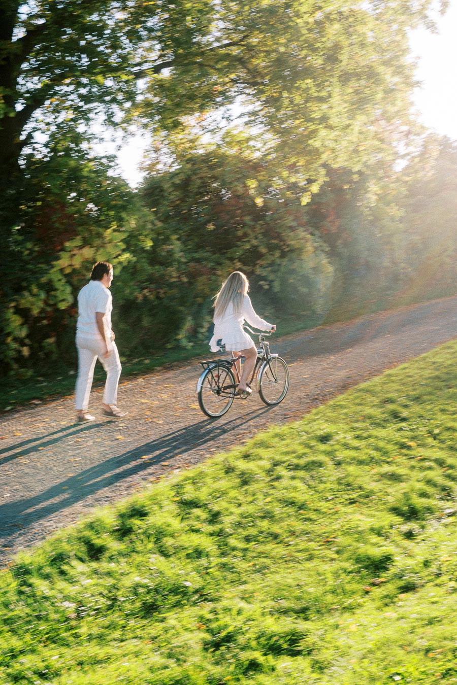 Man and woman in white clothes enjoying a sunny day, with the woman riding a bicycle on a lush green path and the man walking beside her, surrounded by vibrant greenery.