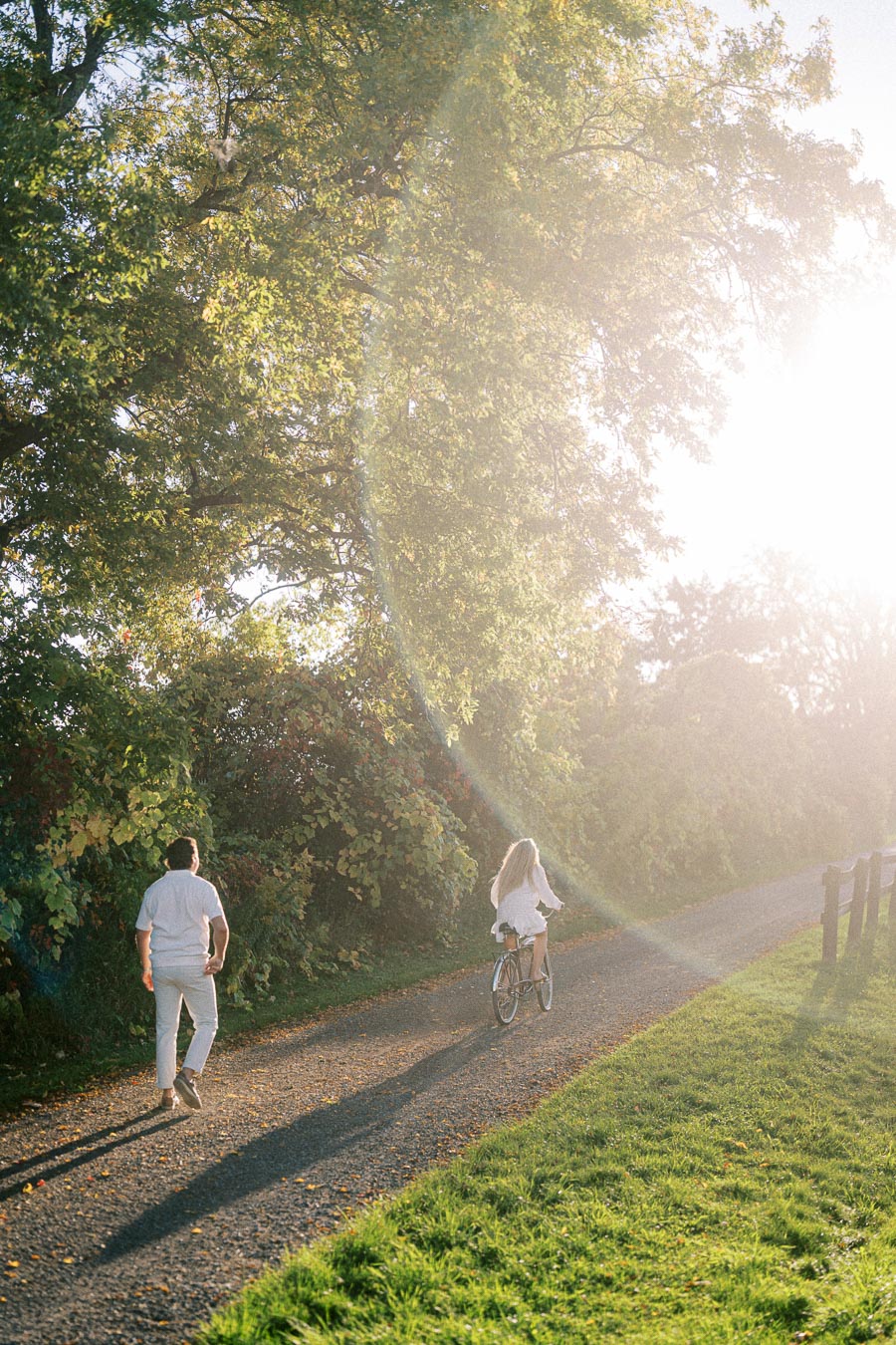 A man and woman enjoying a leisurely walk and bicycle ride on a sunlit tree-lined path in a park.