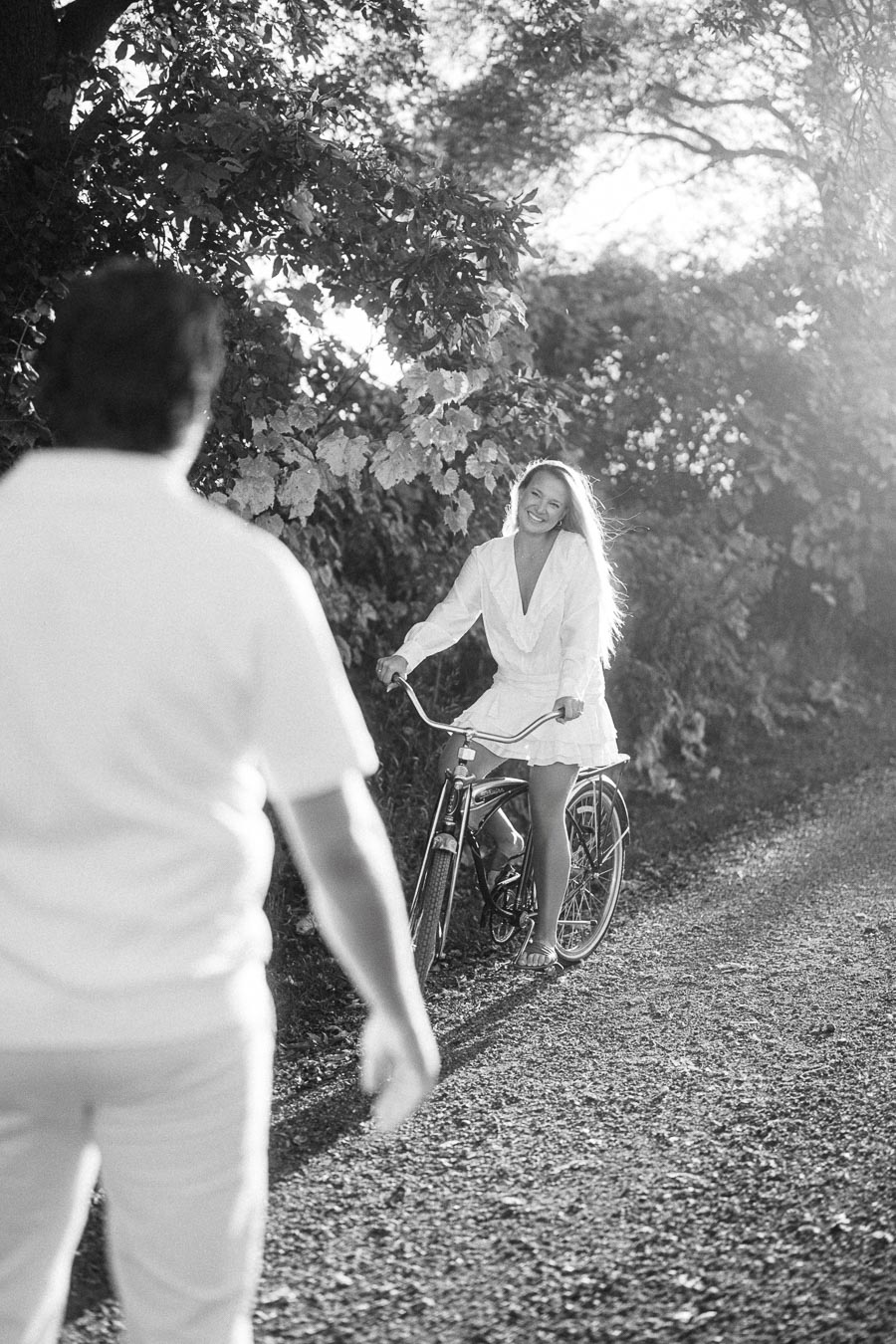 Black and white photo of a woman riding a bicycle on a sunlit path, smiling towards a person walking towards her, with trees in the background.