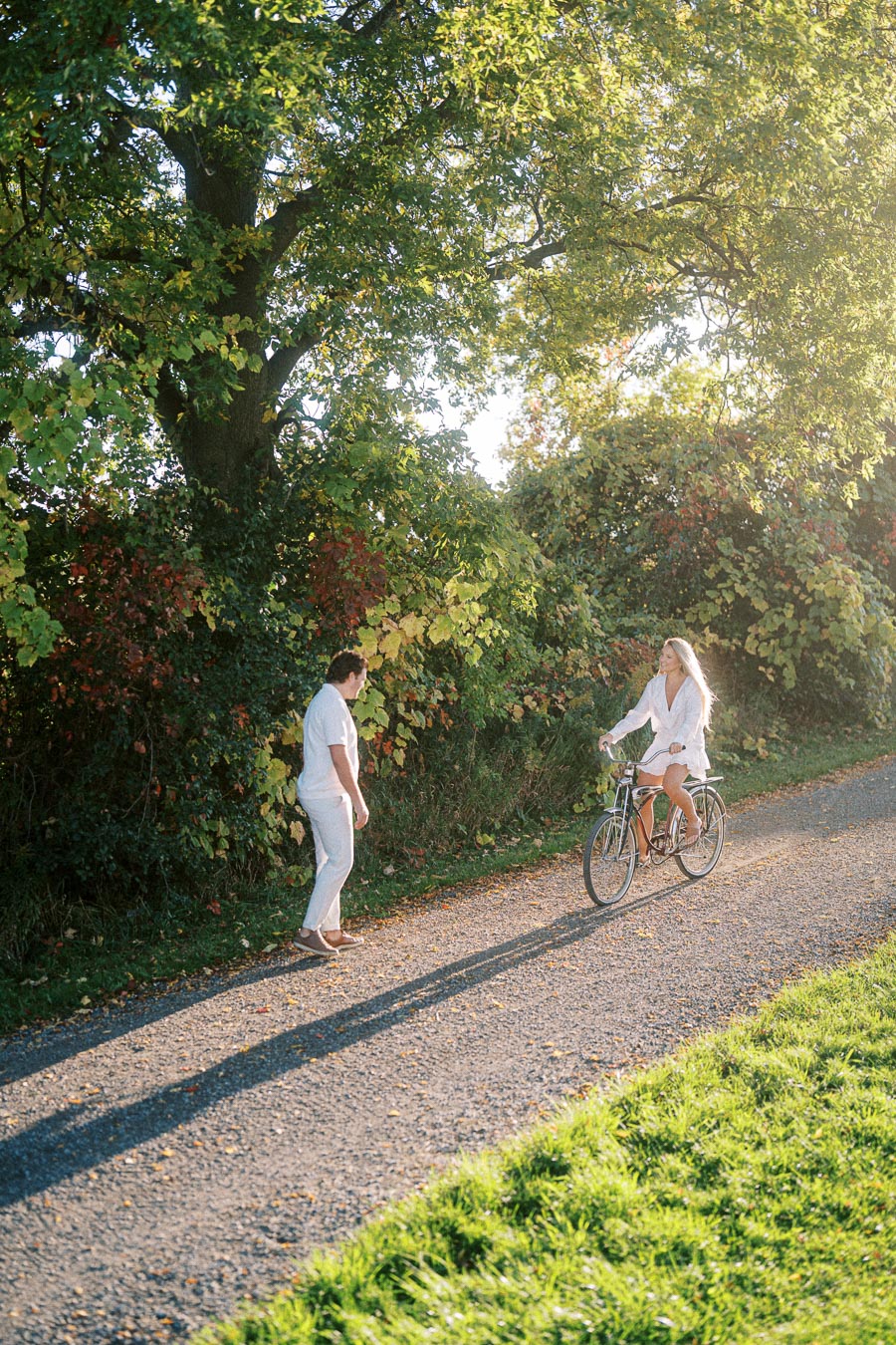 A woman riding a bicycle on a sunlit path through a lush, green park, passing a man walking on the side, surrounded by trees and foliage.