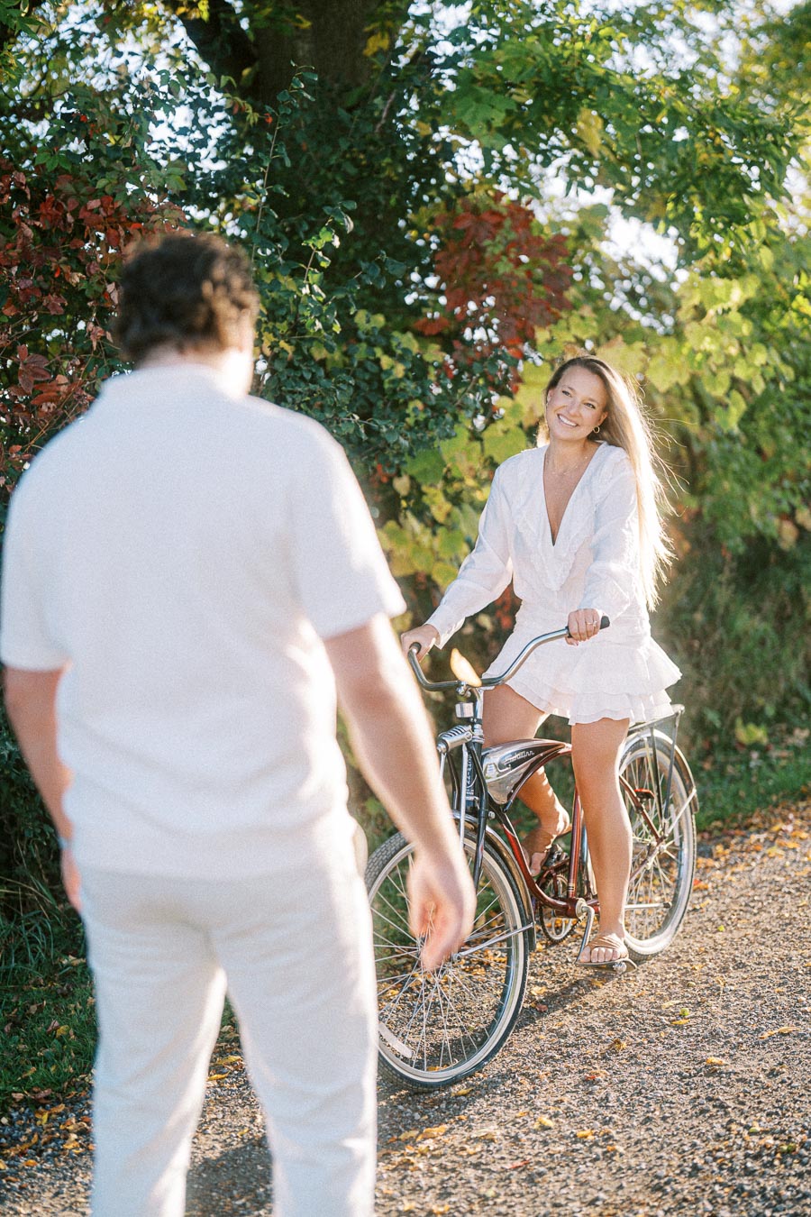 Woman riding a bicycle and smiling on a sunlit path, surrounded by lush green foliage, while a person in white stands nearby.