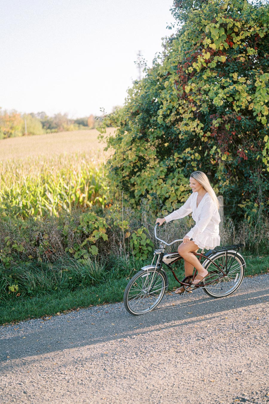 A woman in a white dress riding a vintage bicycle along a rural path surrounded by lush greenery and open fields under a clear blue sky.