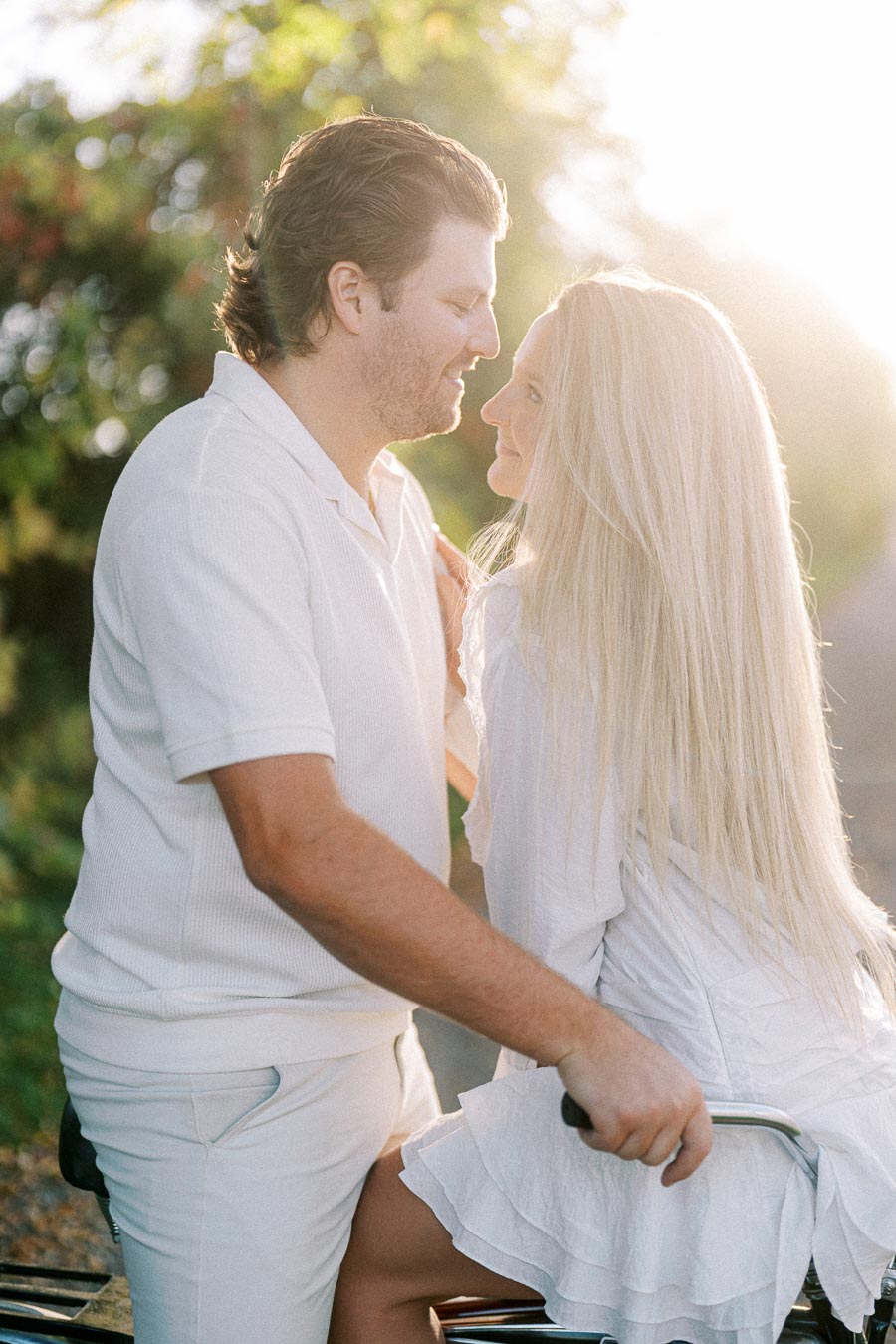 A couple enjoying a romantic moment on a sunny day, with the man sitting on a bicycle and both dressed in white, against a backdrop of lush greenery.