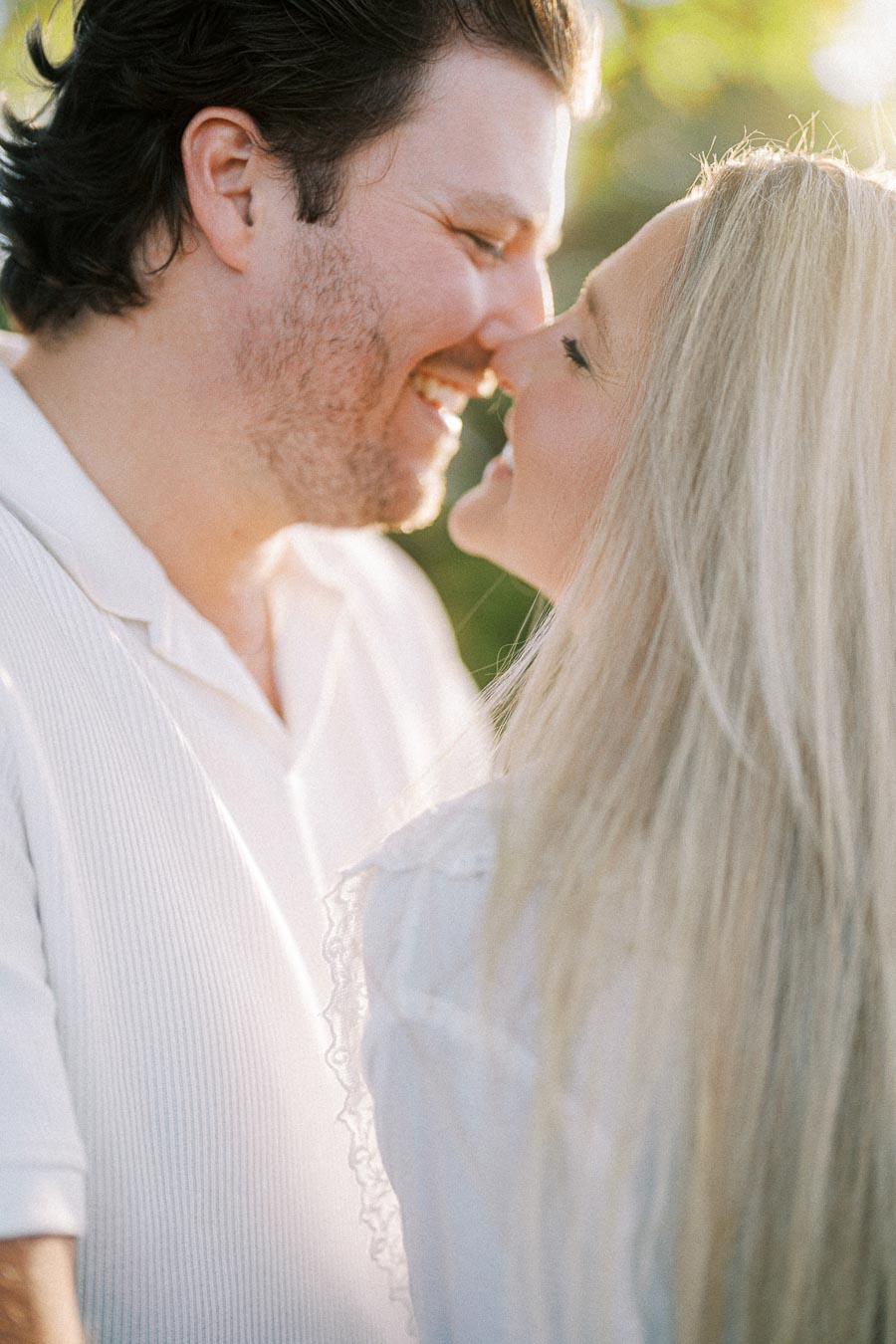 A couple smiling and leaning in for a kiss, surrounded by soft natural lighting and greenery, conveying romance and happiness.