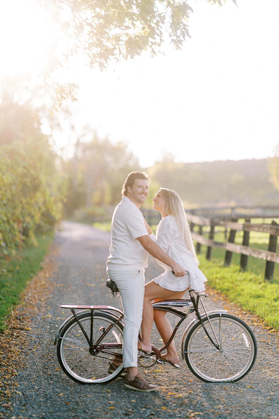 A couple enjoying a romantic moment on a bicycle ride through a sunlit countryside path, surrounded by greenery and a wooden fence.