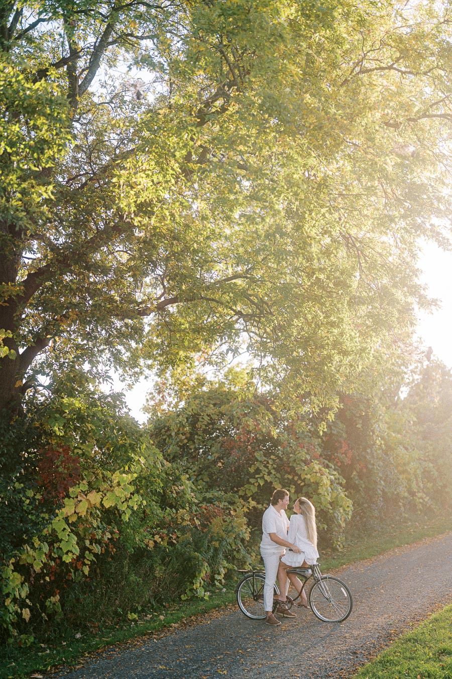 Couple sitting on a bicycle along a scenic path framed by lush green trees bathed in sunlight.
