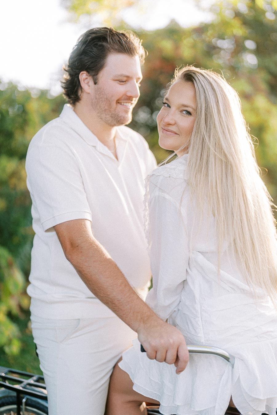 A smiling couple dressed in white, sitting on a bicycle in a sunlit garden.