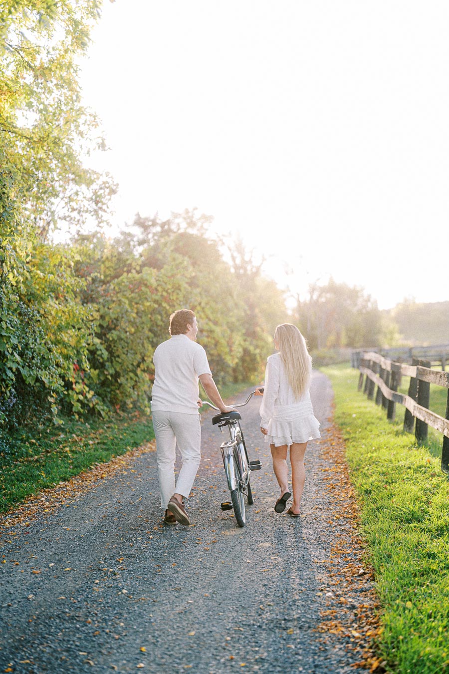 A couple walking with a bicycle on a scenic, sunlit path, surrounded by lush greenery, showcasing a peaceful outdoor setting.