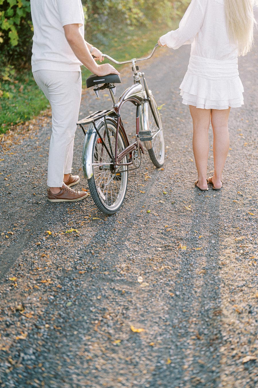 A couple in casual white outfits stands beside a vintage bicycle on a sunlit gravel path, surrounded by green foliage.