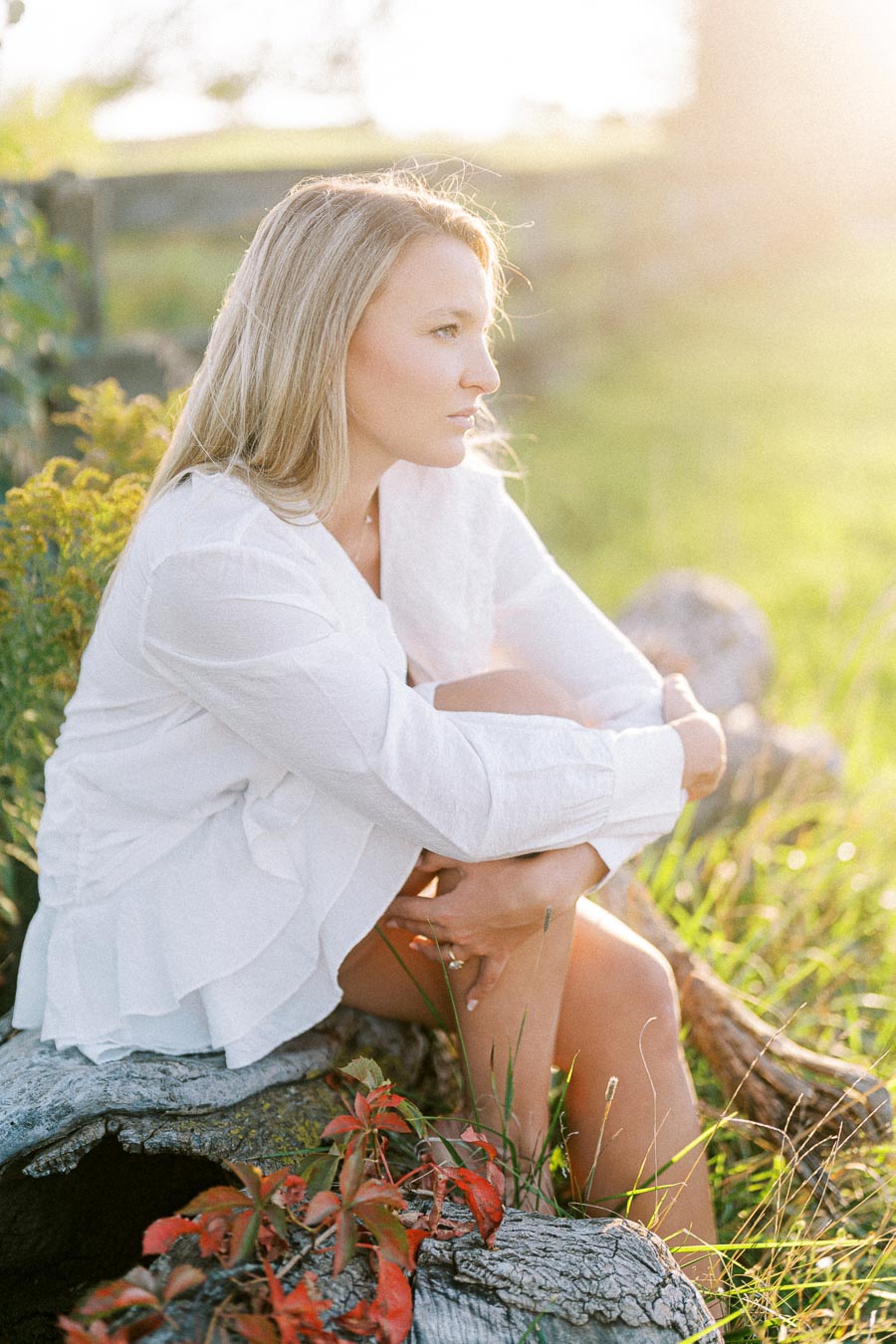 A woman in a white blouse sitting thoughtfully on a log in a sunlit outdoor setting, surrounded by green grass and red leaves, with a wooden fence in the background.