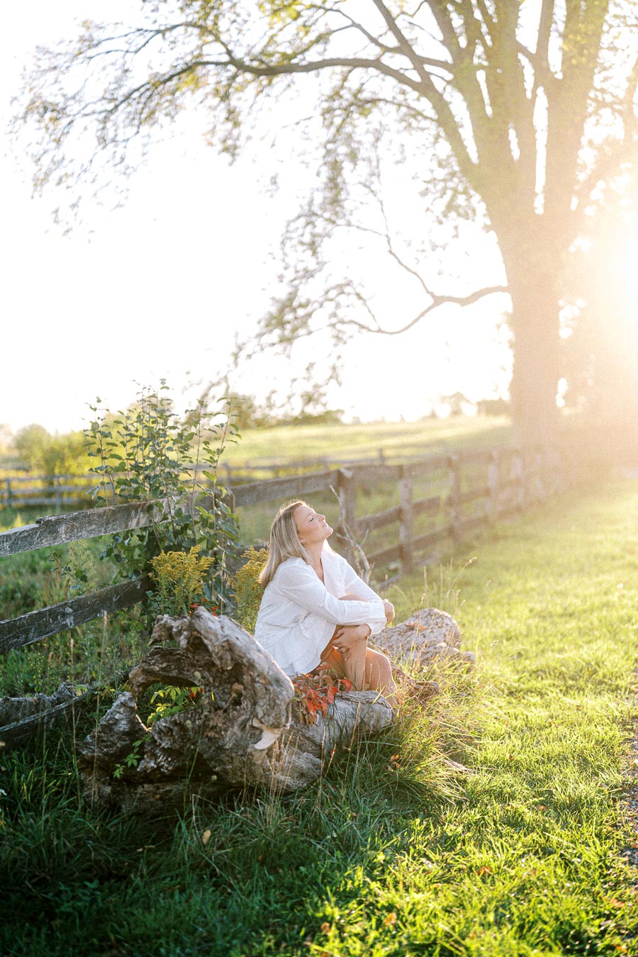 Woman sitting on a fallen log in a sunny meadow, surrounded by lush greenery and a wooden fence. She is enjoying the warm sunlight and the natural, serene environment.