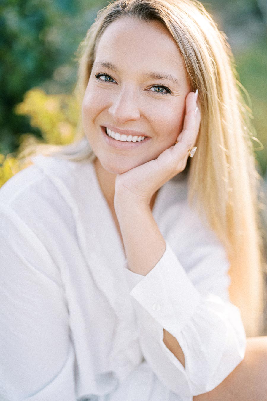 Portrait of a woman with long blonde hair smiling in a white blouse, set against a blurred natural background.