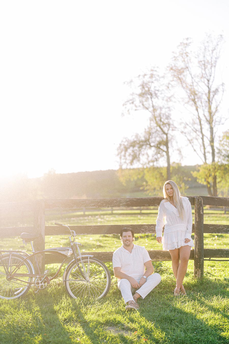 A couple in casual white attire enjoying a sunny day by a rustic wooden fence with a vintage bicycle, surrounded by lush green grass and trees.
