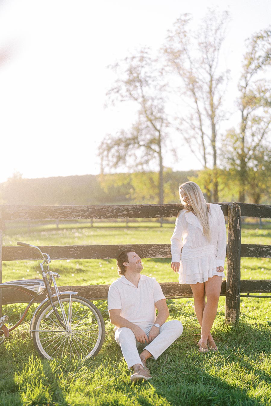 Couple enjoying a sunny day in a rural setting, with a vintage bicycle and wooden fence in the background. The woman stands smiling while the man sits on the grass, both dressed in casual white clothing.