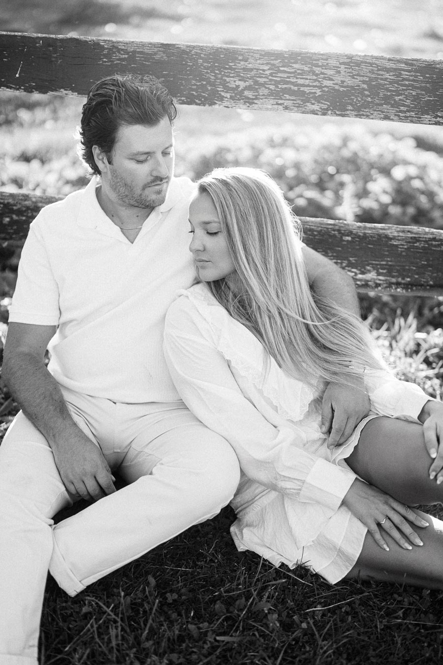 A black and white image of a couple sitting closely on the grass, leaning against a wooden fence, bathed in soft sunlight. The man is wearing a white shirt and pants, while the woman is in a light dress, both with serene expressions, symbolizing tranquility and connection outdoors.