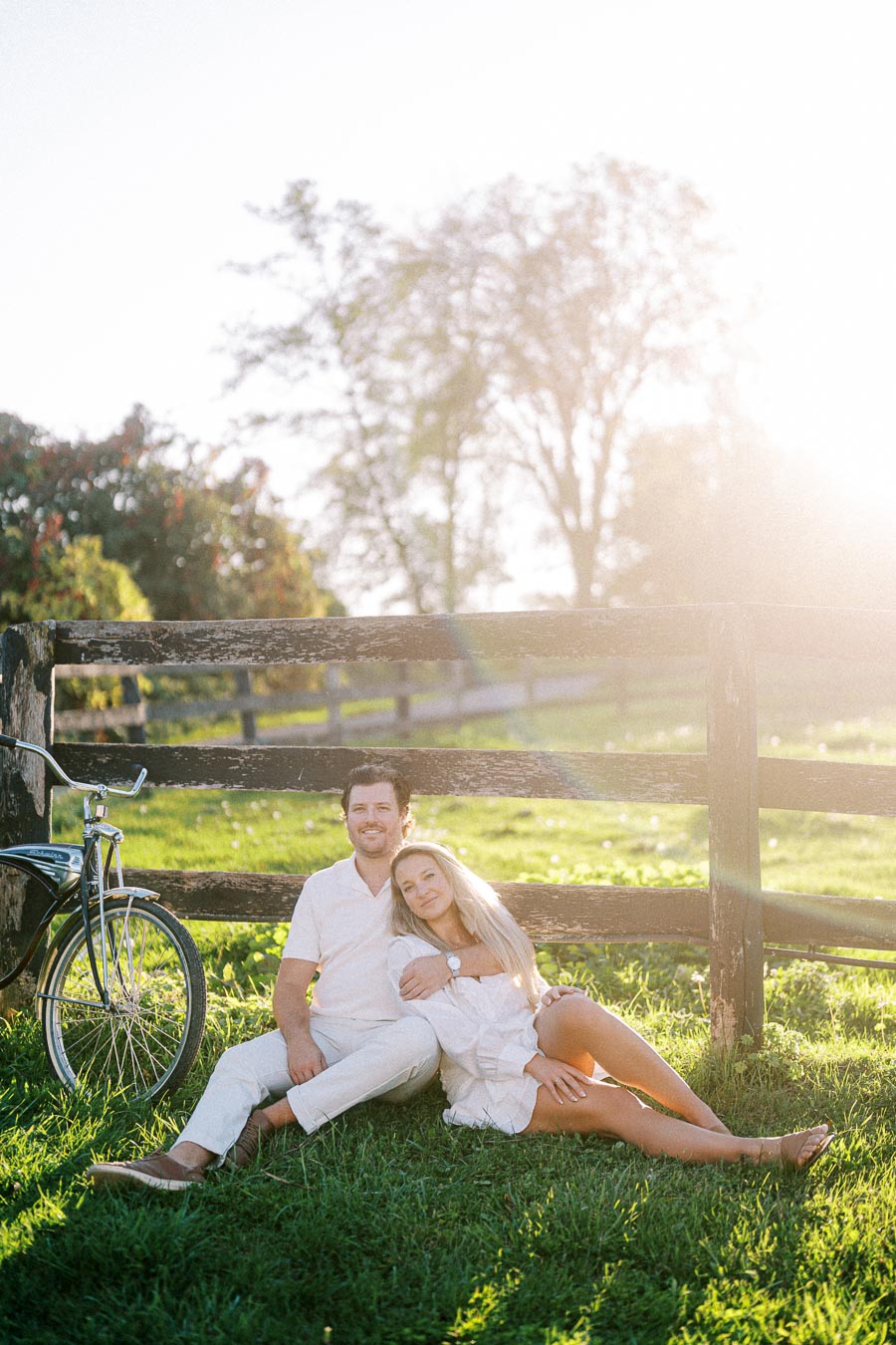 A couple relaxing in a sunlit field, leaning against a wooden fence with an old-fashioned bicycle nearby, trees in the background, capturing a serene and romantic outdoor moment.