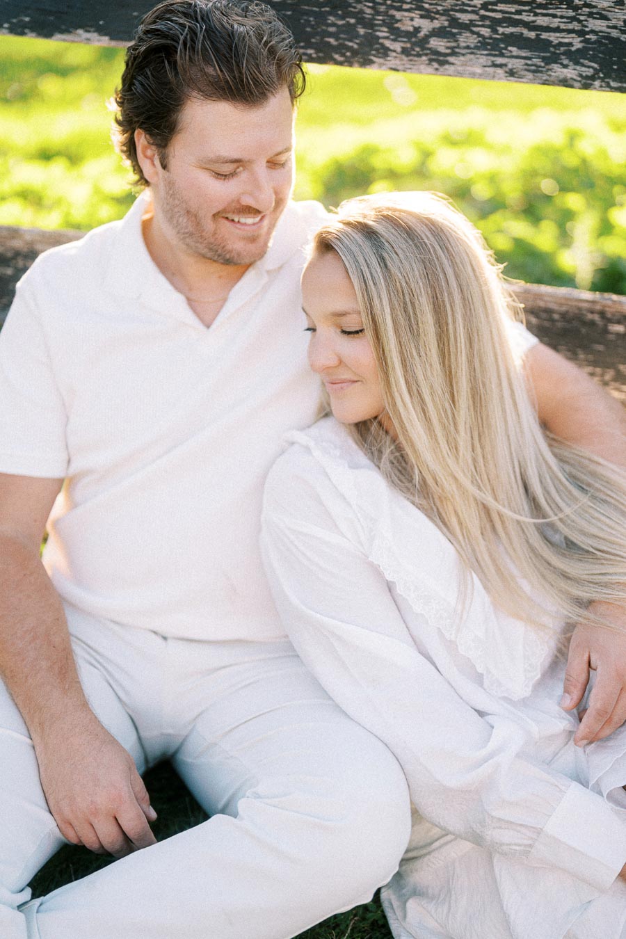 A couple sitting on grass, wearing matching white outfits, smiling and embracing each other in a serene outdoor setting with lush green background.