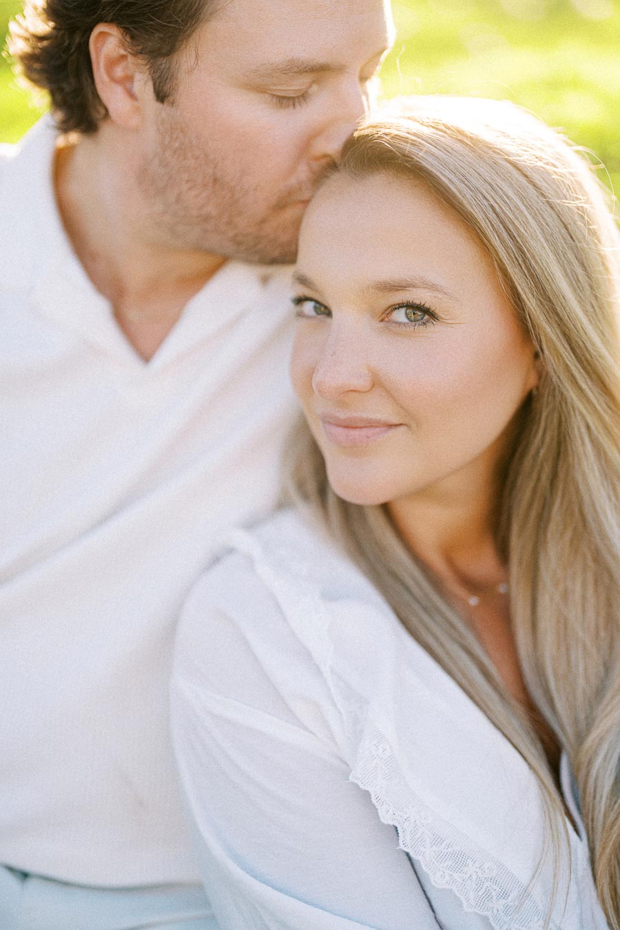 A couple in a sunlit setting, with a man gently kissing a woman's forehead, both wearing white clothing, conveying love and tenderness.