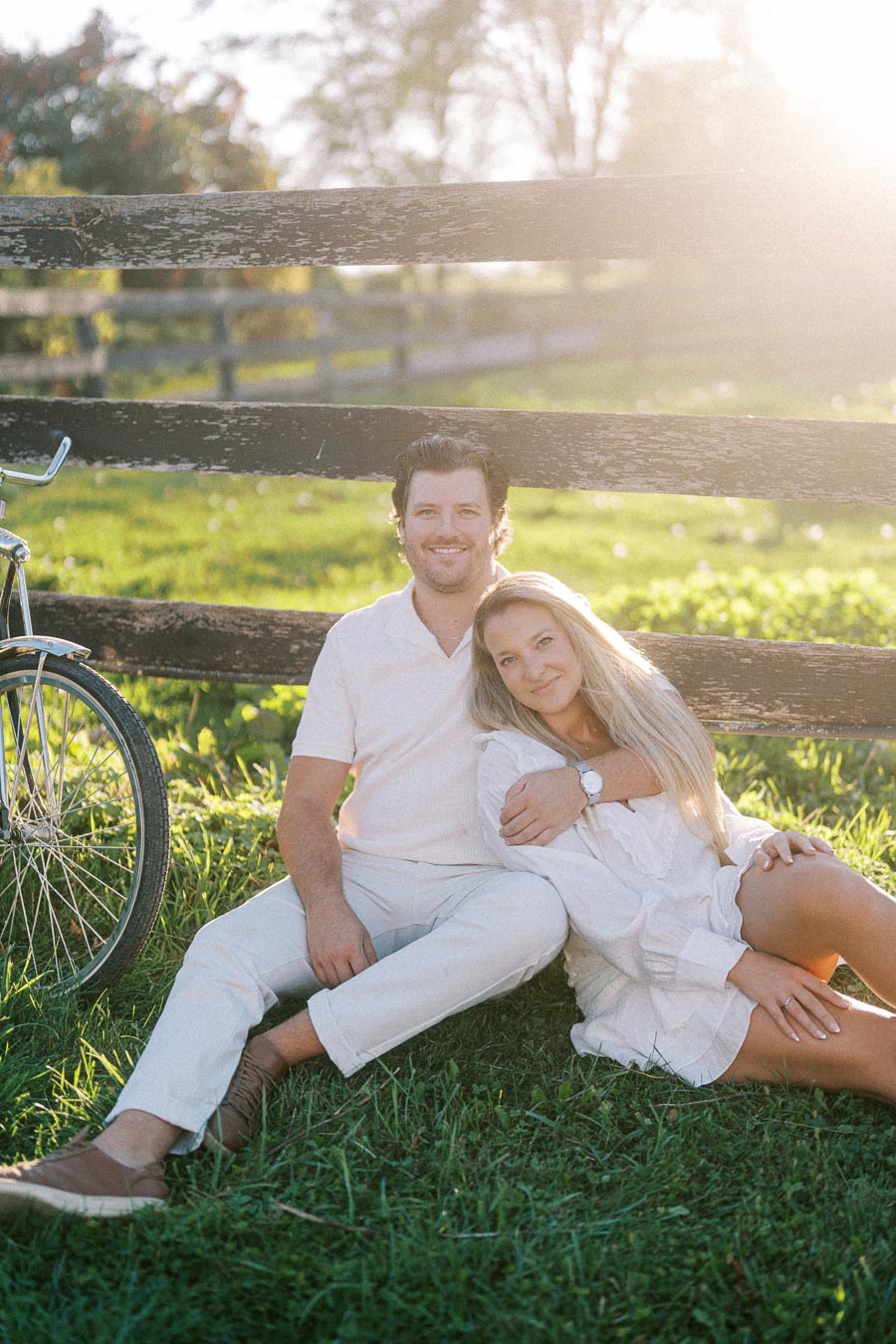 A couple sitting on grass near a rustic fence, with a vintage bicycle nearby, enjoying a sunny day in a serene countryside setting.