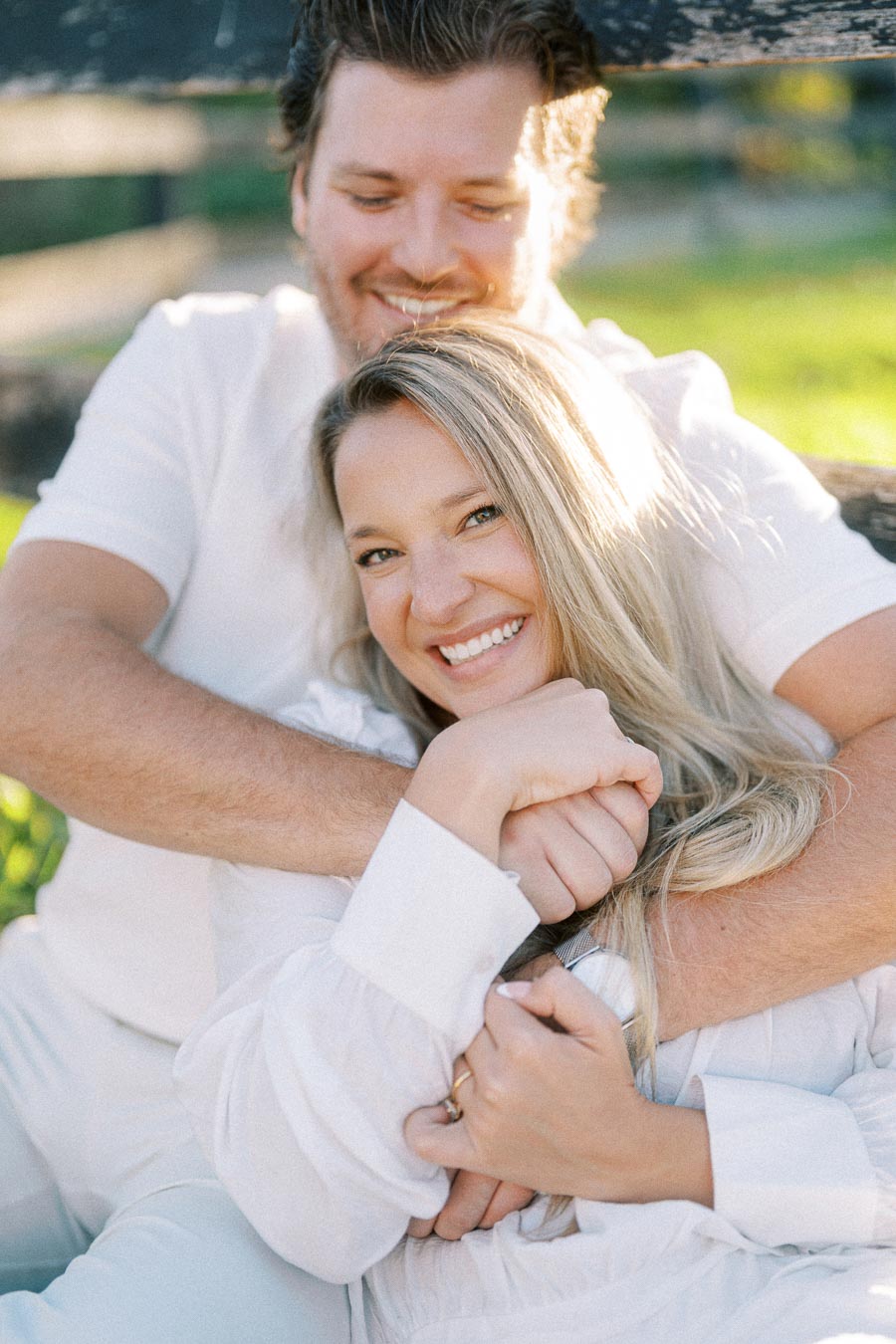 A couple embracing and smiling in a sunlit outdoor setting, with greenery in the background, conveying warmth and happiness.