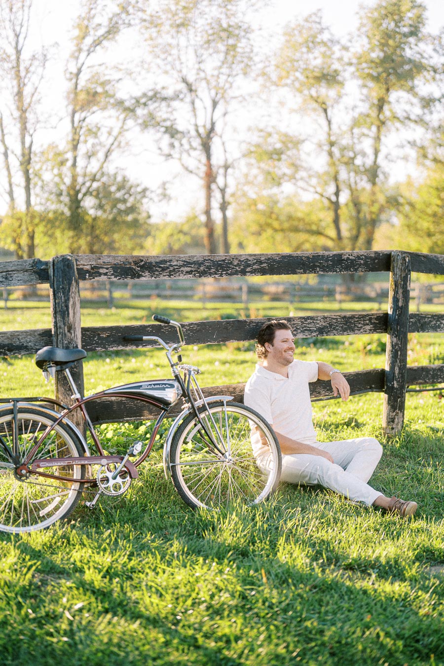 Man sitting on grass beside a vintage bicycle and wooden fence, enjoying a sunny day in a grassy field with trees in the background.