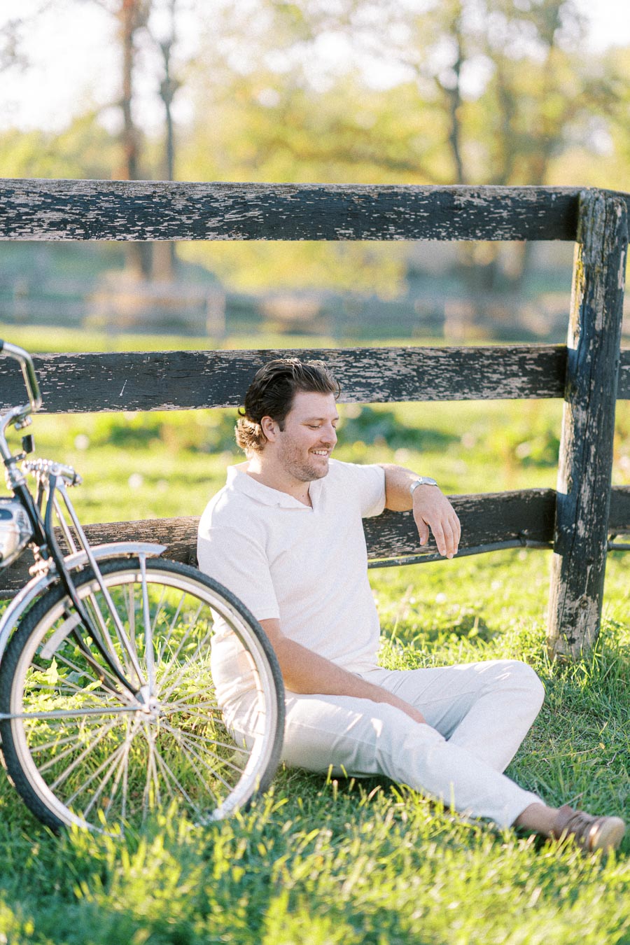 Man sitting on grass by a wooden fence, next to a bicycle, enjoying a sunny day outdoors.