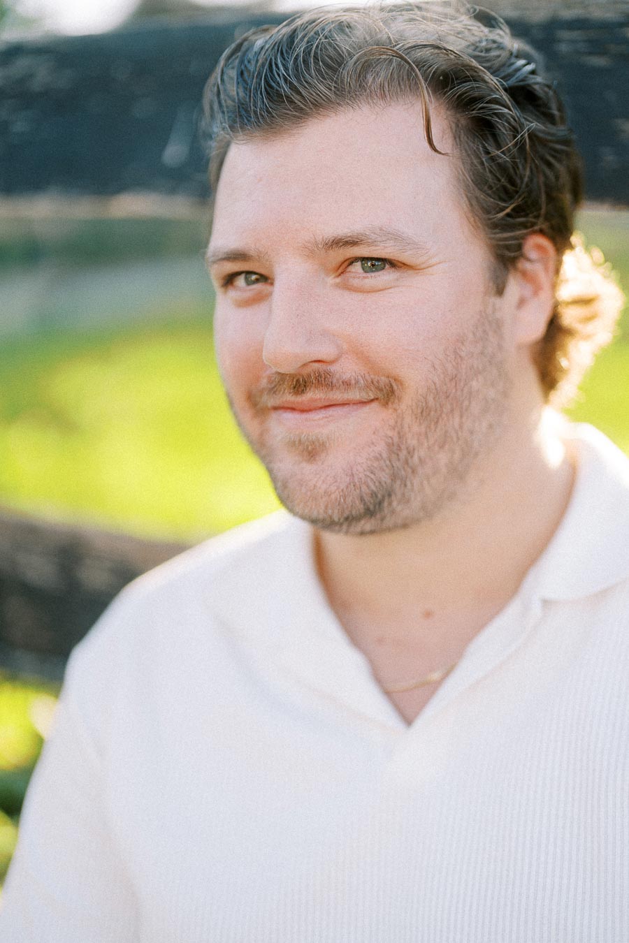 A man with short brown hair and a beard smiling, wearing a white shirt, standing outdoors with a blurred green background.