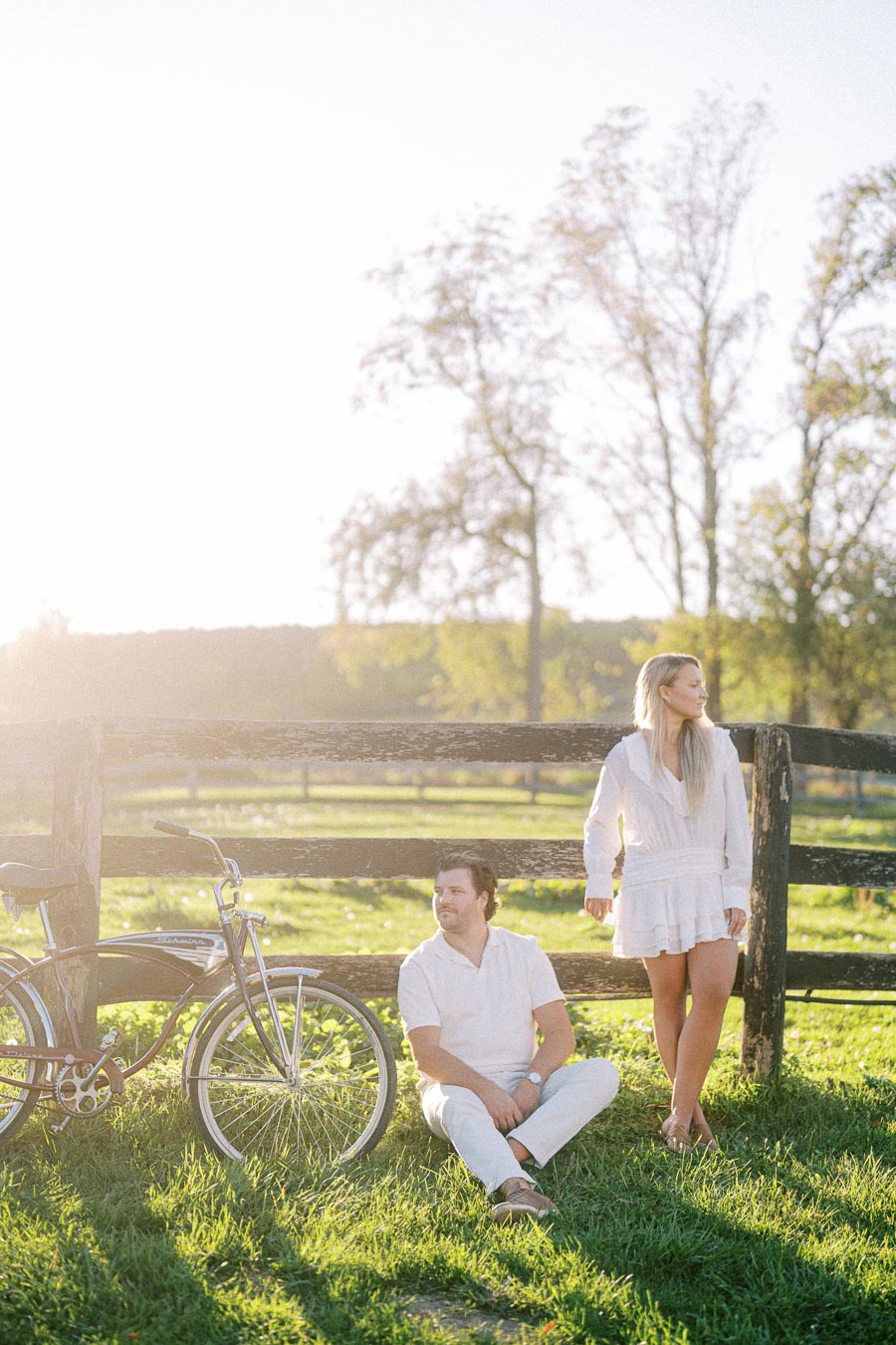 Couple enjoying a sunny day in a countryside setting, with a vintage bicycle leaning against a rustic wooden fence and tall trees in the background, highlighting a serene outdoor atmosphere.