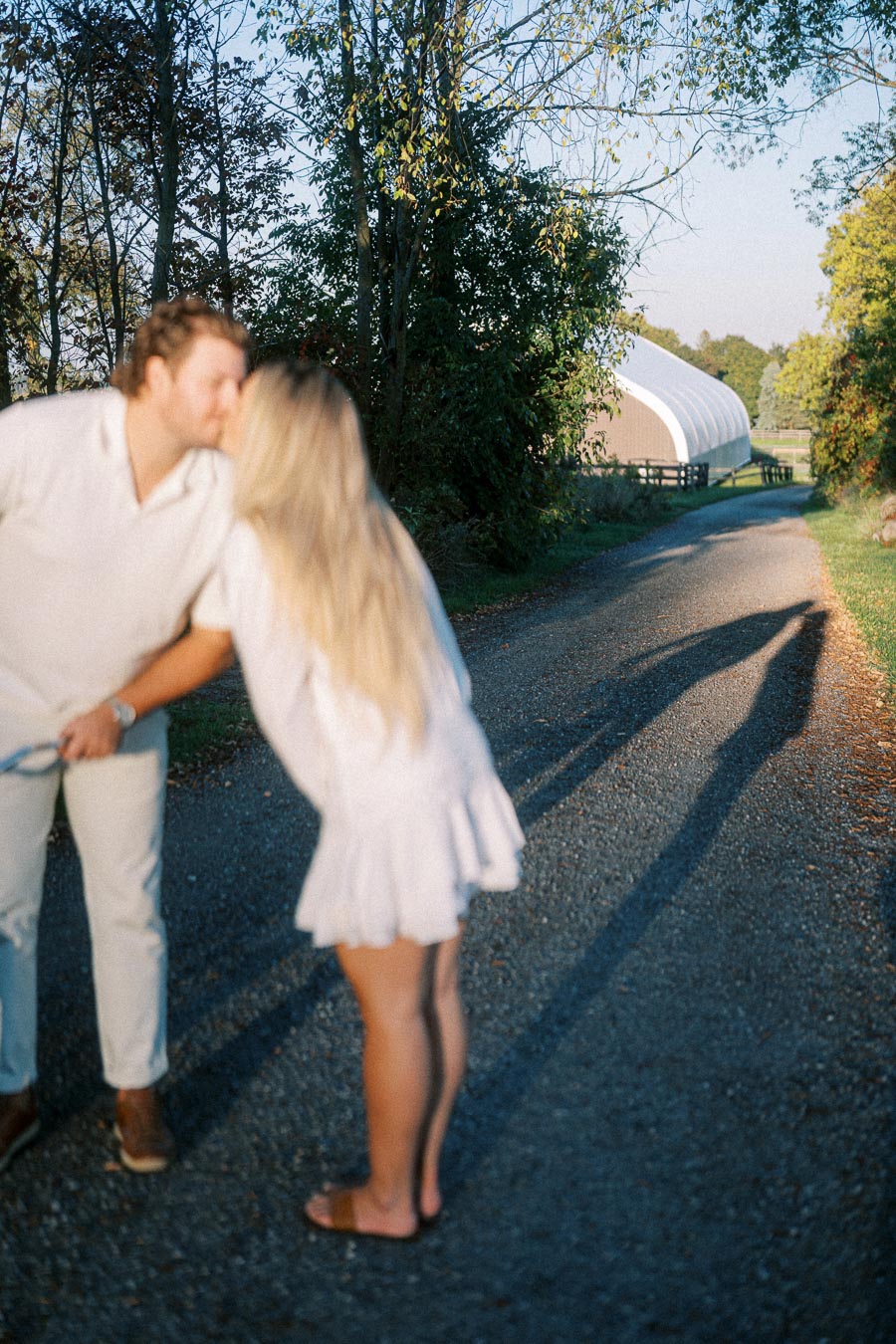 A couple in casual white outfits shares a kiss on a country road lined with trees, with a barn and lush greenery in the background on a sunny day.