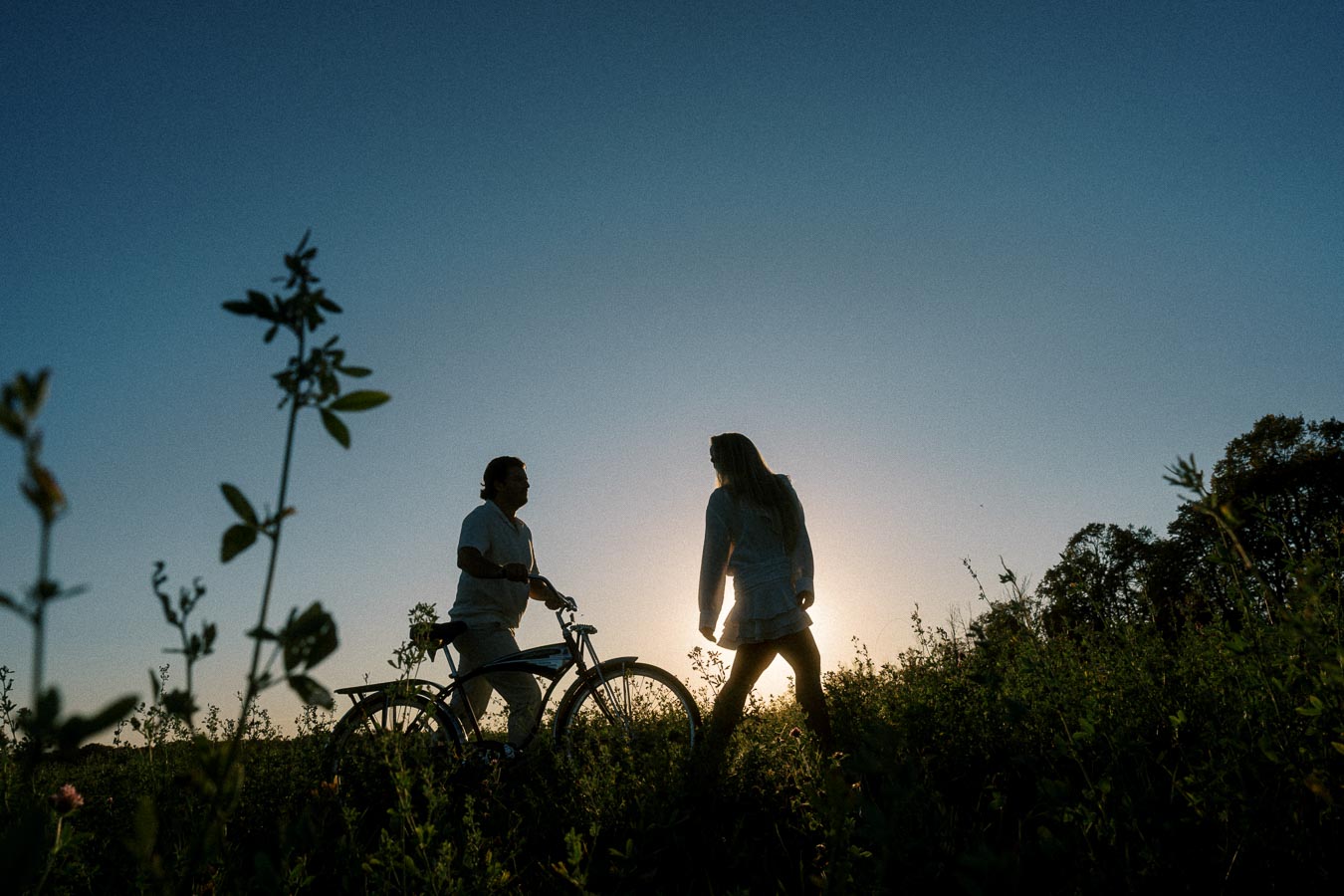 A silhouette of two people enjoying sunset, with one person standing next to a bicycle and the other walking through a field, surrounded by nature and a clear sky.