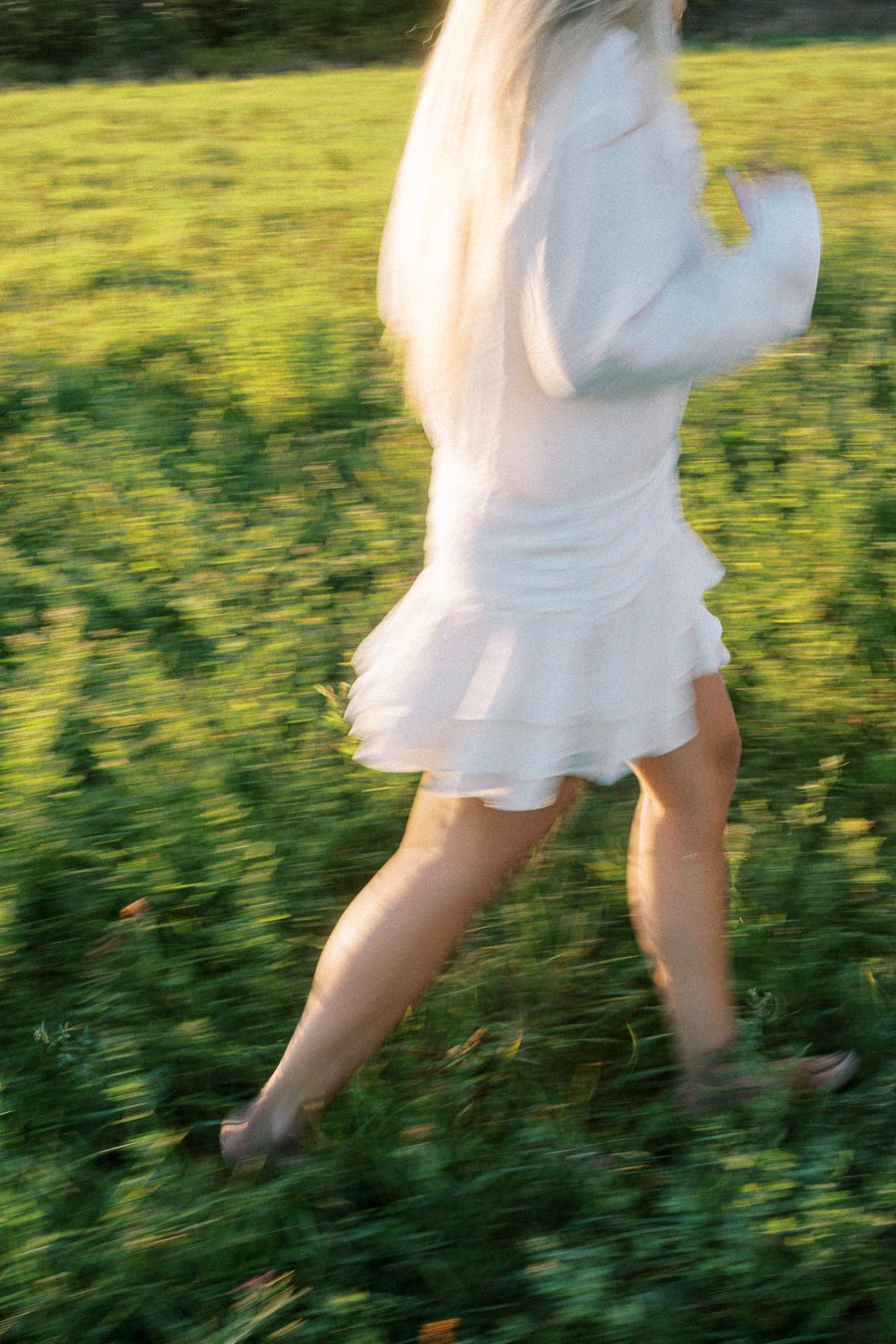 Blurred image of a woman with long hair in a white dress, running through a sunlit green field, capturing a sense of movement and freedom.