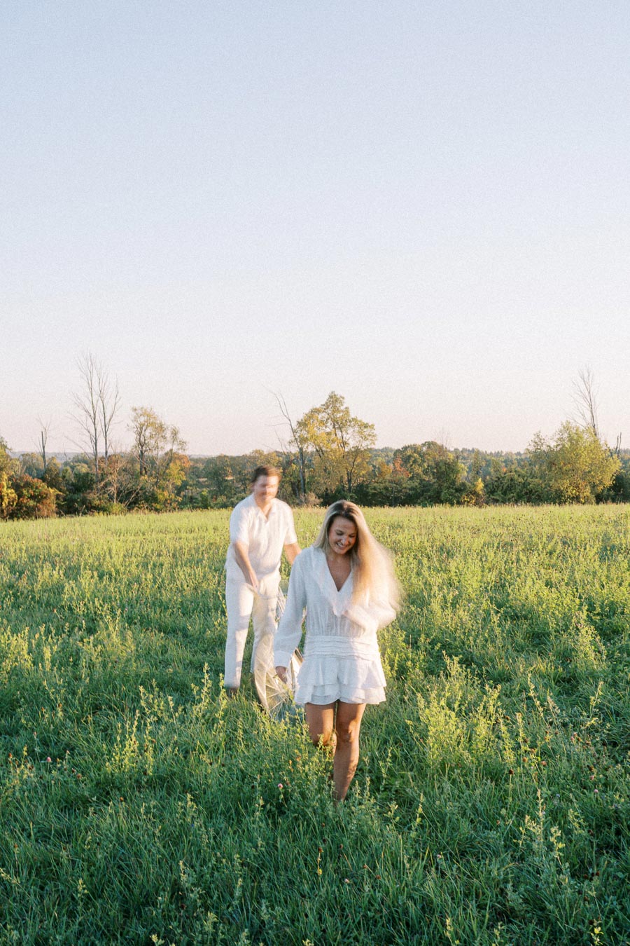 A couple in white clothing walking joyfully through a sunlit green meadow, with a clear blue sky in the background, capturing a serene and happy moment in nature.