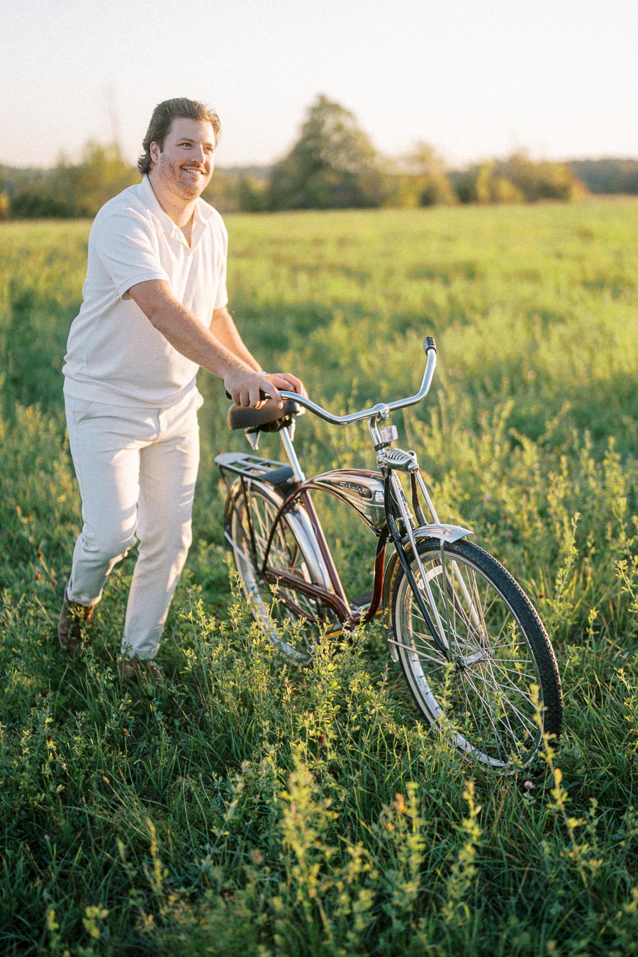 A person in a white outfit walks with a vintage bicycle through a sunlit green field, enjoying a peaceful outdoor moment.