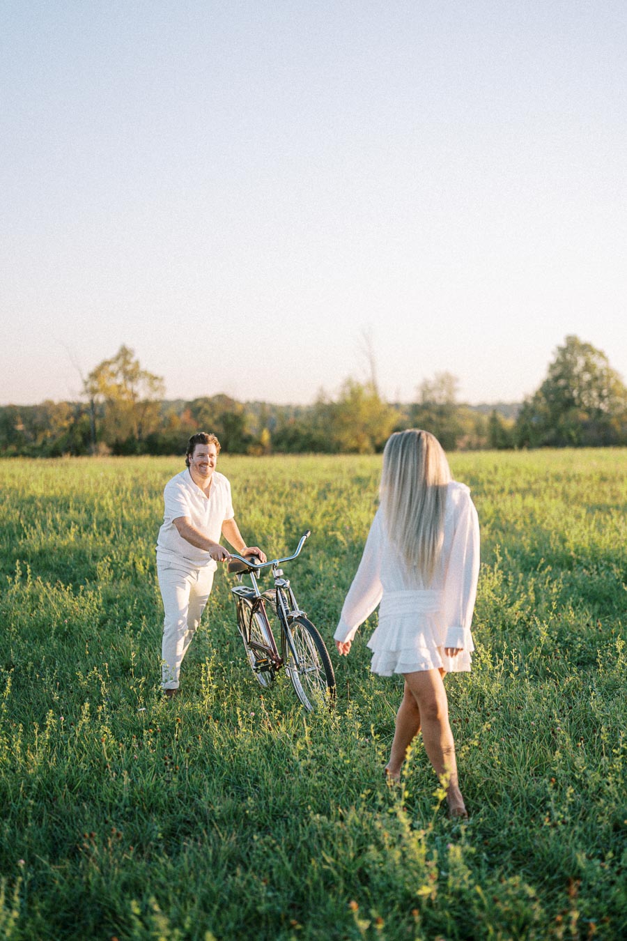 A couple in a grassy field enjoys a sunny day; a man with a bicycle approaches a woman wearing a white dress, framed by lush greenery and a clear blue sky.
