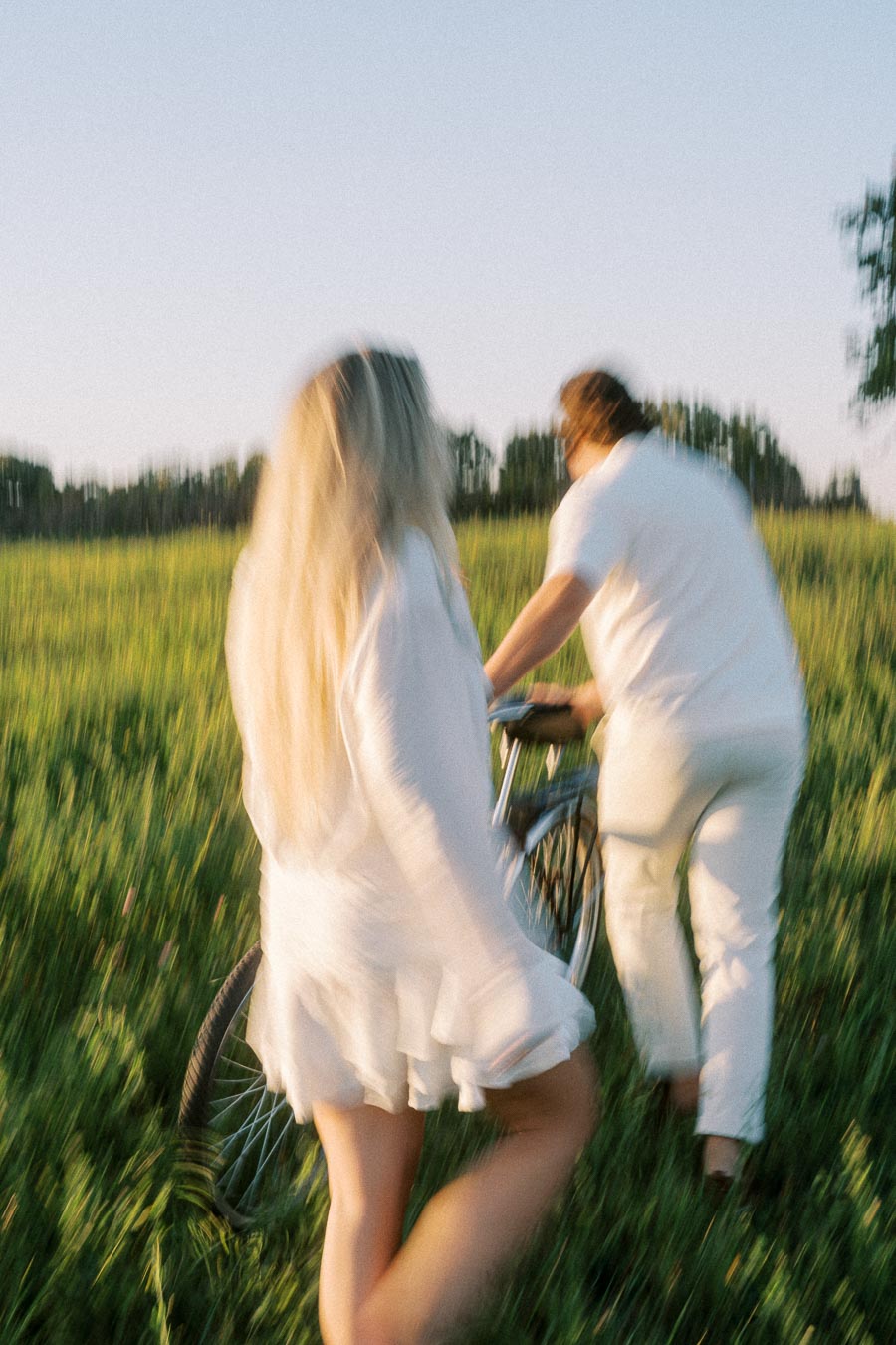 Blurry image of two people walking with a bicycle through a grassy field during sunset, wearing light-colored clothing.