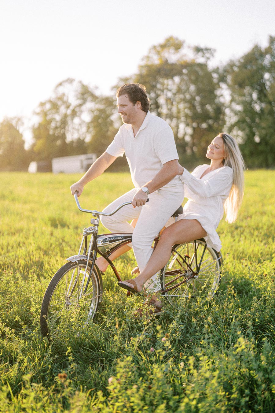 A couple enjoying a tandem bicycle ride in a sunny green field, wearing casual white clothing, with trees in the background and a warm, joyful atmosphere.