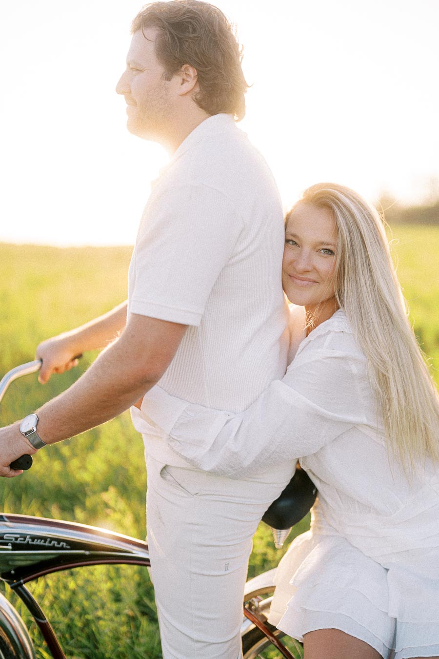 A couple dressed in white, enjoying a sunny day outdoors; the woman smiles and embraces the man as they stand next to a vintage bicycle in a lush green field.