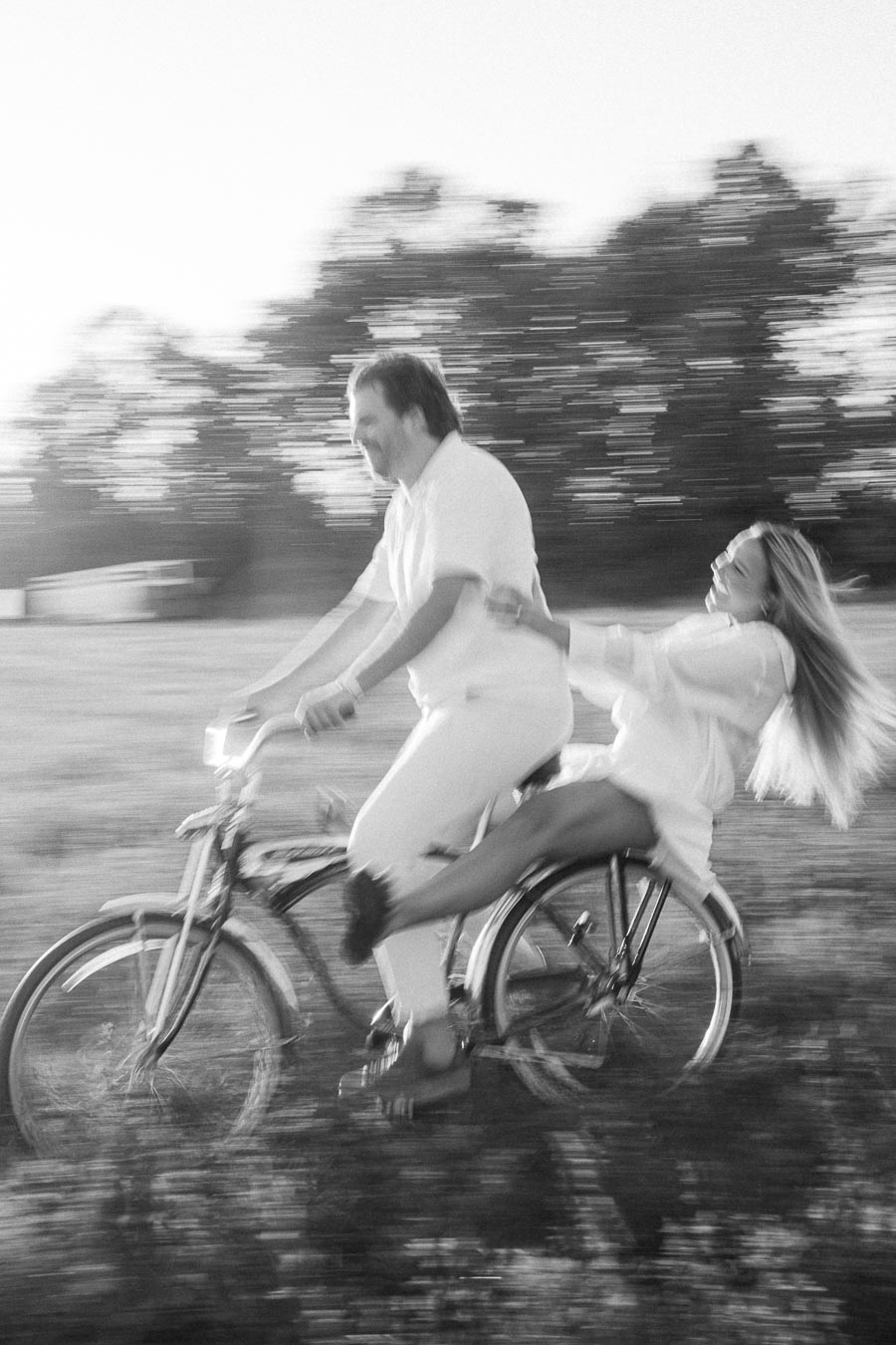 Black and white image of a couple joyfully riding a bicycle through a field, with the man pedaling and the woman laughing as she sits on the back, surrounded by blurred motion suggesting speed and movement, conveying a sense of freedom and happiness.