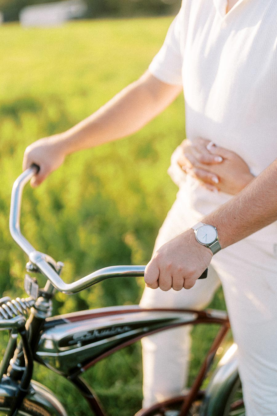 A person dressed in white casually holds onto bicycle handlebars, while another person gently embraces them from behind, set against a vibrant green field backdrop, exuding a sense of warmth and companionship.