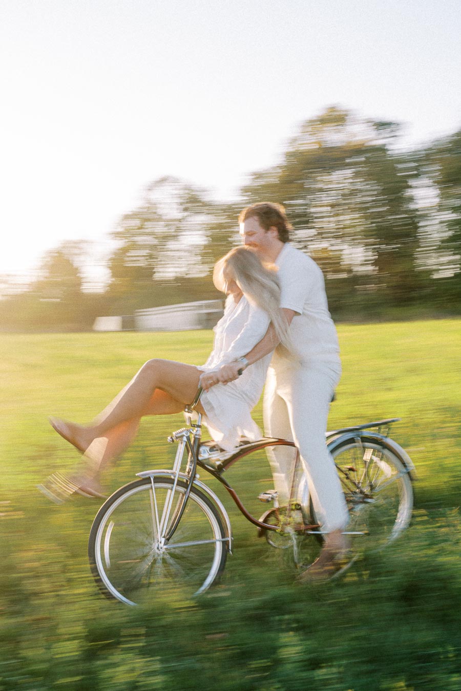 A couple joyfully riding a bicycle together in a sunny green field, capturing a playful and carefree moment with motion blur.