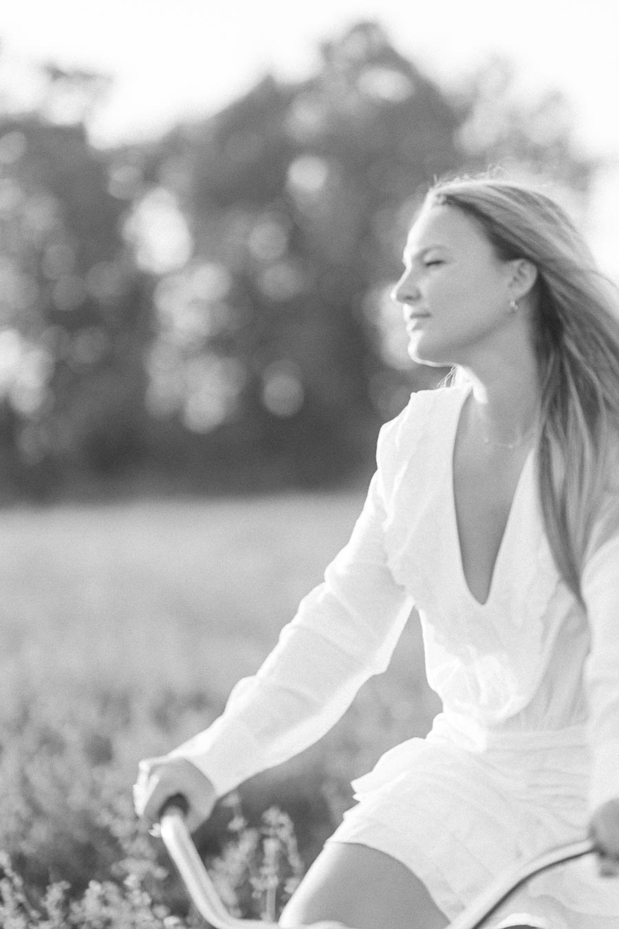 Young woman in a white dress riding a bicycle through a sunny field, with blurred trees in the background, enjoying a peaceful outdoor moment.