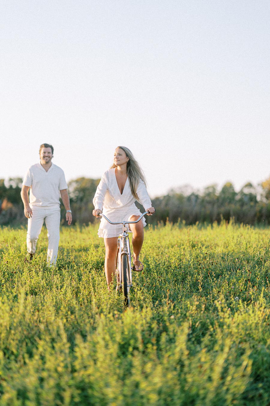 A woman in a white dress rides a bicycle through a sunny green field, while a man walks behind her, both enjoying a peaceful outdoor moment.