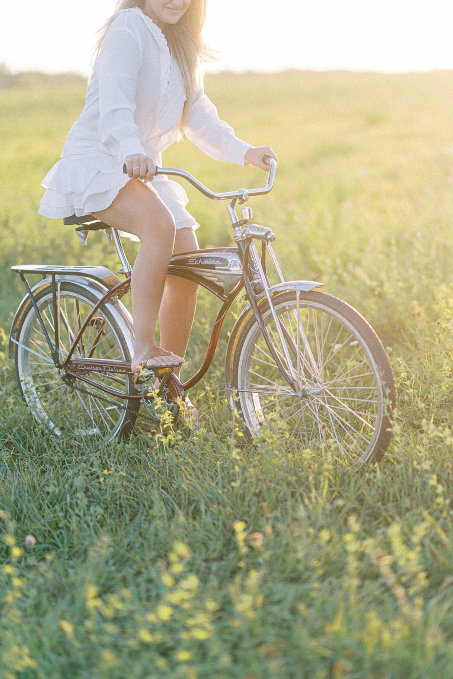 A woman rides a vintage Schwinn bicycle through a sunlit, grassy field, wearing a white dress, capturing a serene and carefree summer moment.