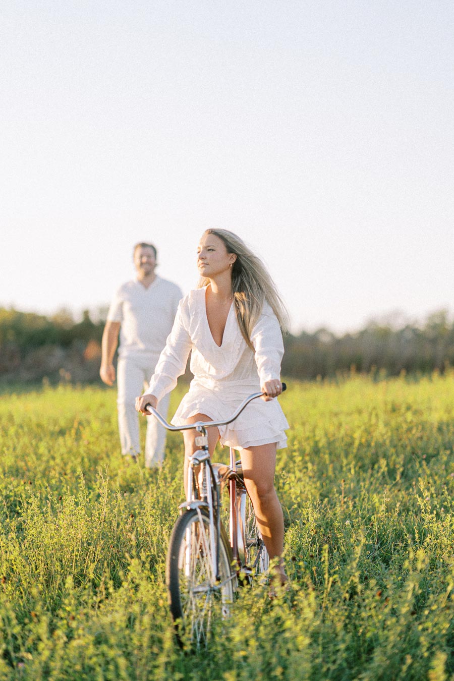Woman in a white dress riding a bicycle through a green field with a man walking behind her under a clear blue sky.