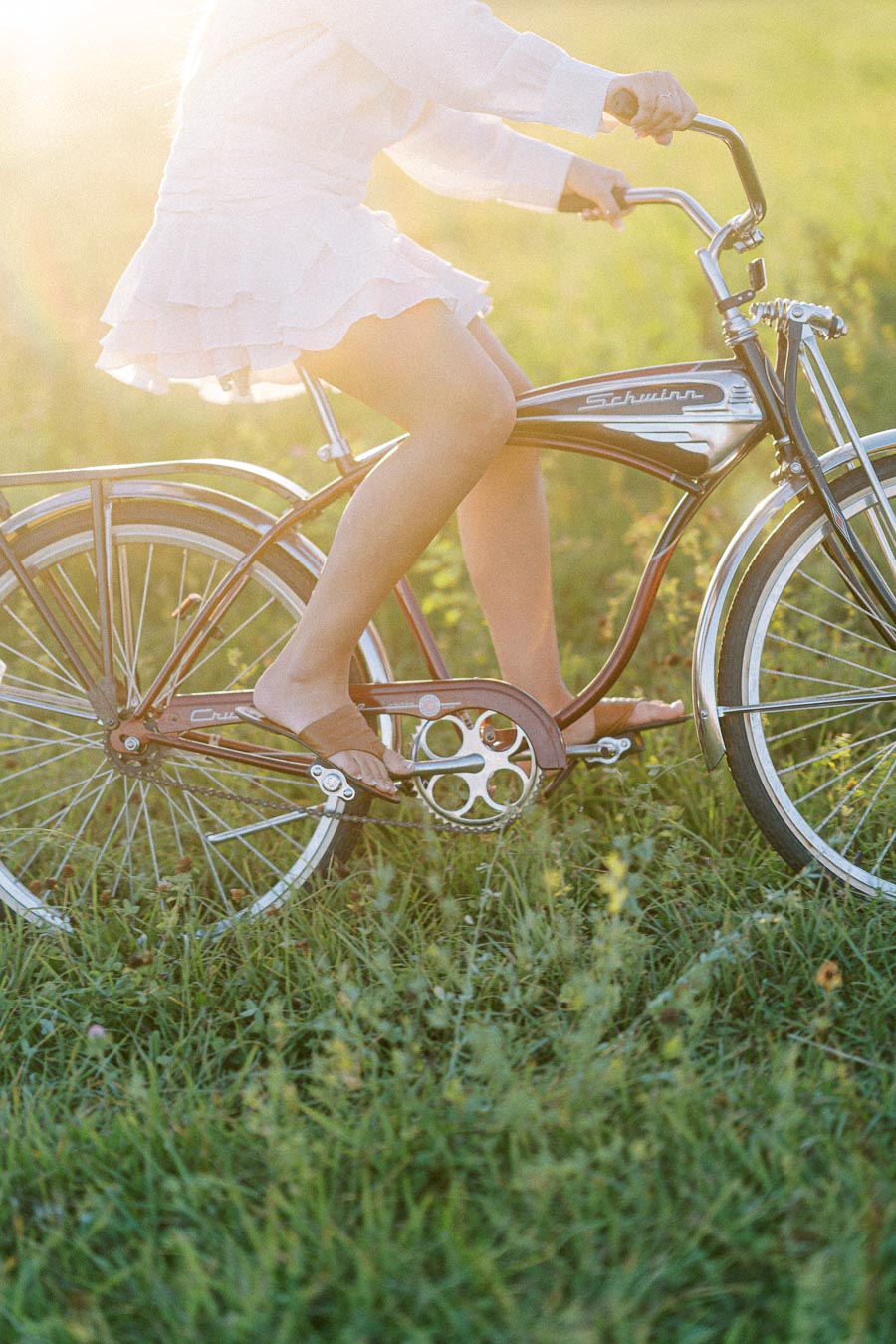 A person riding a vintage bicycle in a sunlit grassy field, wearing a white dress and sandals, capturing a peaceful and nostalgic summer moment.