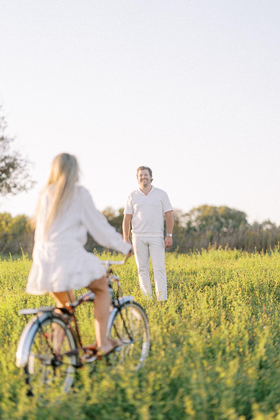 A woman in a white dress riding a bicycle towards a man in a white outfit standing in a lush green meadow under a clear sky.