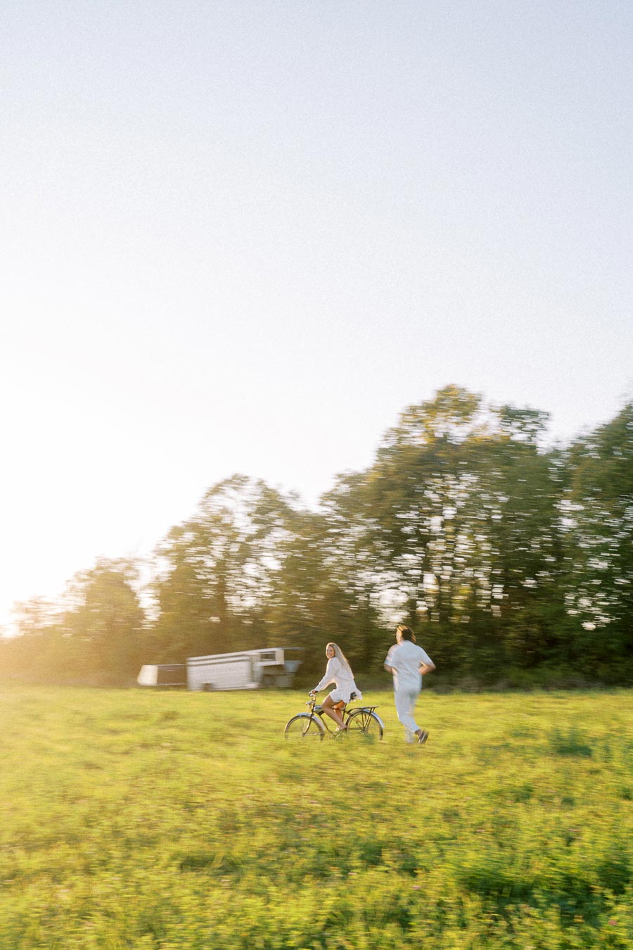 A person cycling through a sunny field with another person running beside, amidst lush greenery and a clear blue sky.
