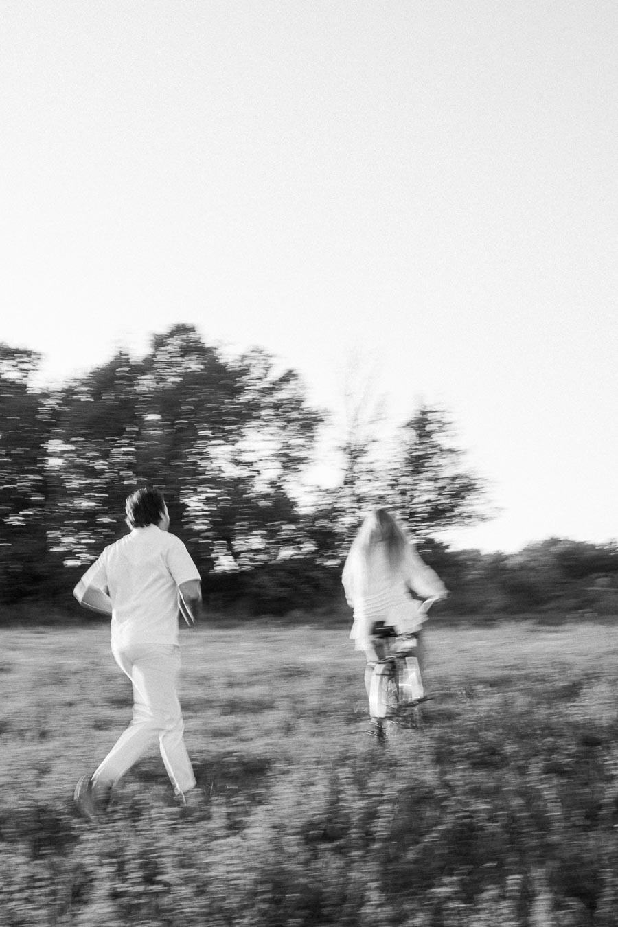Blurred black and white photo of a person running and another riding a bicycle in a grassy field with trees in the background.