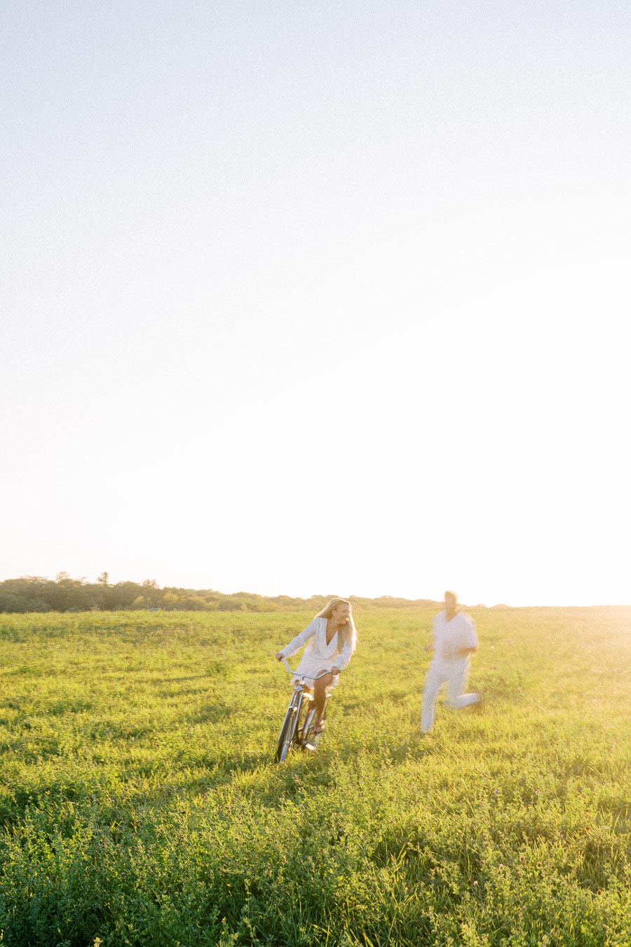 A person riding a bike through a sunlit, grassy field while another person runs behind, capturing a carefree summer moment outdoors.